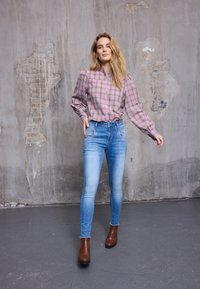Camicia a quadri rosa e verde, maniche a palloncino, jeans a vita alta azzurri e stivaletti marroni su uno sfondo grigio strutturato.