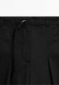 Gros plan d’un pantalon noir avec une ceinture en tissu assortie passée dans des passants et fermée avec une boucle en D argentée.