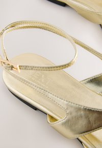 Sandali metallici dorati con sottile cinturino alla caviglia, soletta testurizzata e suola piatta nera. Presentano il logo impresso sulla parte interna del plantare.