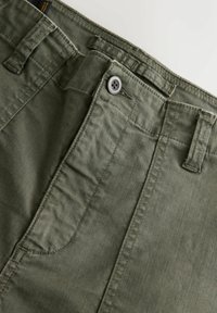 Close-up of green pants showing buttoned waistband, belt loops, and top portion of front pocket on a light background.
