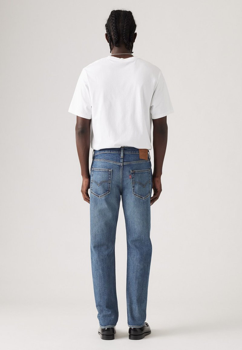 Vue de dos d'une personne portant un t-shirt blanc uni et un jean droit bleu avec deux poches arrière et un patch en cuir.