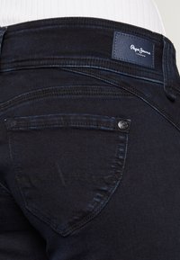 Tmavě modré denimové džíny s zadní kapsou s jemným prošíváním a s označením značky. Tkanina vypadá hladce s mírným leskem.