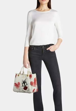 Donna che indossa una maglia bianca a maniche lunghe e jeans neri tiene in mano una borsa bianca con il disegno di un gatto fumettistico e cuori rossi.