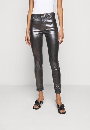 Femme portant un pantalon skinny noir brillant, un top blanc sans manches et des sandales à talons noires avec des brides nouées, debout sur un fond uni.
