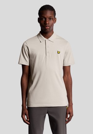 Beige polo skjorta med krage, korta ärmar och tre knappar. Har en liten gul fjäril-logotyp på vänster bröst.
