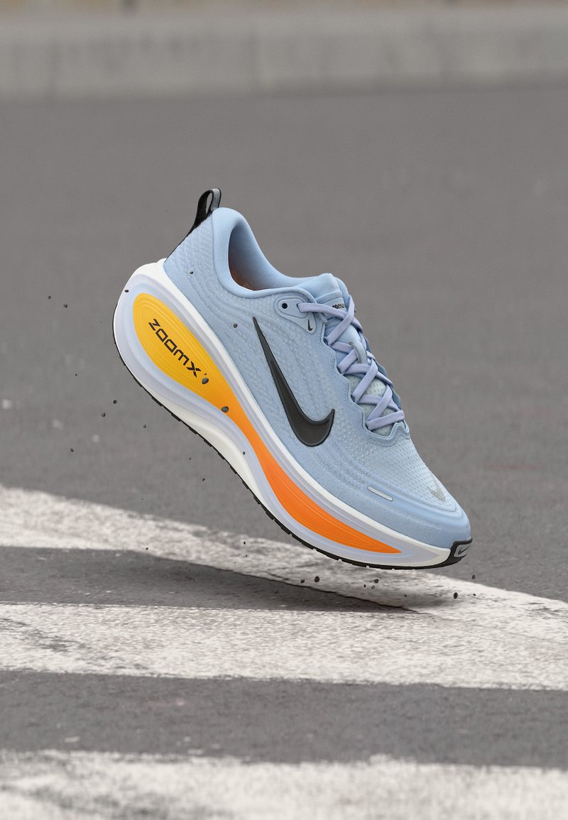 Υποδήματα Nike για τρέξιμο σε ανοιχτό γαλάζιο χρώμα με μαύρο λογότυπο και πορτοκαλί σόλα ZoomX, φωτογραφημένα στον αέρα πάνω από διάβαση πεζών σε άσφαλτο.