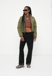Giacca puffer cropped verde oliva con un piccolo emblema, top corto marrone, pantaloni larghi neri e sneakers grigie; modella con occhiali da sole.