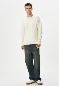 Cremefarbener Pullover mit Rundhalsausschnitt, kombiniert mit dunkelblauen Jeans. Das Model trägt weiße und braune Sneaker. Glatte Textur.