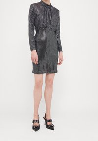 Robe noire en sequins avec des manches longues, un col plissé, une taille ajustée et une longueur au genou. Assortie à des escarpins noirs à bout pointu avec des lanières.