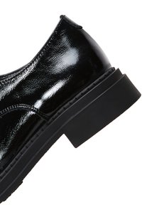 Schwarzer, polierter Lederschuh mit glatter Textur, runder Spitze und dicker schwarzer Sohle, die eine leichte Höhenanhebung aufweist.