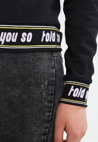 Sudadera negra con puños acanalados en contraste que presenta el texto "te lo dije" en blanco, bordeado por una franja amarilla. Falda de mezclilla visible.