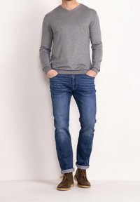 Pull gris à manches longues avec col en V, jean en denim bleu avec légère décoloration, et chaussures marron à lacets avec une semelle robuste, posant devant un fond neutre.