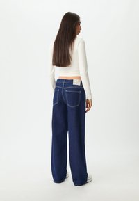 Široke tamnoplave traperice s bijelim šavovima i oznakom na stražnjem dijelu struka. Model nosi usku bijelu majicu dugih rukava.