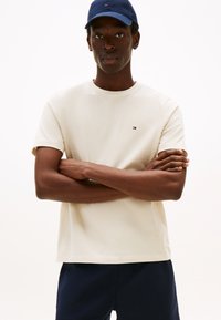 Beežist puuvillast lühikeste varrukatega ja ümar kaelusega T-särk, millel on rinnal väike logo punase, valge ja sinise värviga.