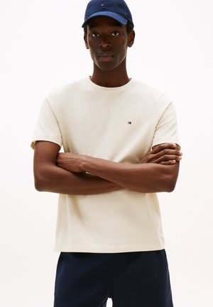 Beige bomuld t-shirt med korte ærmer og rund halsudskæring, med en lille logo i farverne rød, hvid og blå på brystet.