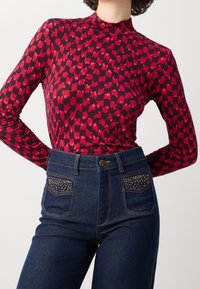 Maglietta a maniche lunghe con un motivo astratto rosso e rosa scuro, abbinata a jeans in denim scuro a vita alta con applicazioni di borchie sulle tasche.