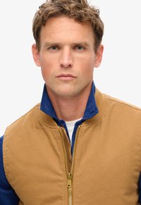 Veste bomber zippée beige avec un col bleu ; fabriquée en tissu lisse avec une finition texturée ; dotée d'une fermeture éclair dorée et de poignets côtelés.