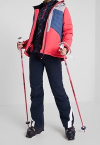 Ski-Outfit mit einer mehrfarbigen, wattierten Jacke mit blauen und roten Einsätzen, einem marineblauen Oberteil, schwarzen Hosen mit weißen Streifen und pinkfarbenen Skistöcken.