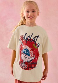 Mosolygó, szőke hajú gyermek, aki krémszínű, túlméretezett pólót visel, melyen színes fagylalt-parfé mintája látható, rózsaszín háttér előtt.