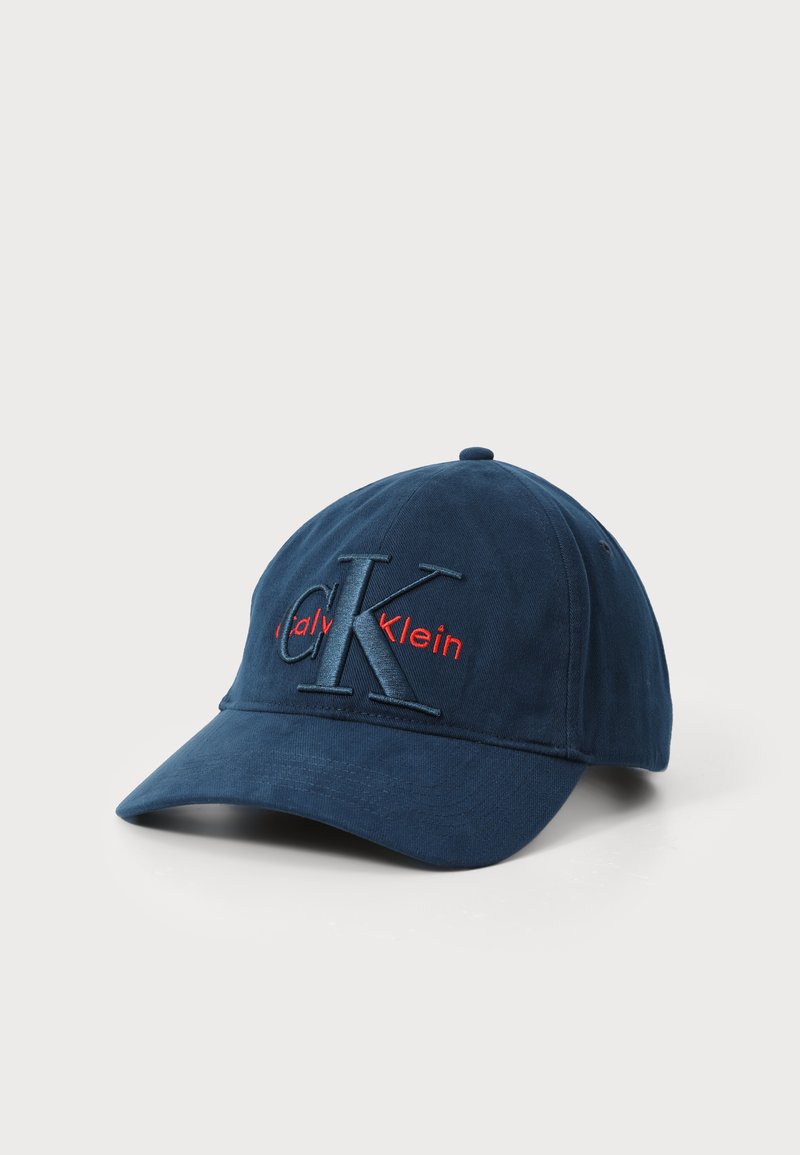 Marineblaues Baseballcap mit gebogenem Schirm, versehen mit gesticktem "CK"-Logo in dunklen und roten Fäden. Hergestellt aus weichem Stoff mit strukturiertem Finish.