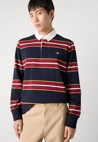Marineblå langermet polo med røde, gule og hvite horisontale striper, med hvit krage og en liten brodert logo på brystet.