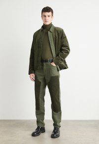Military green corduroy jacket over a matching turtleneck and pants, featuring textured patches. Black belt and shoes complete the outfit.