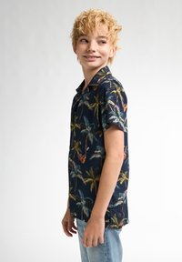 Cămașă pe care scurtă, de culoare bleumarin, cu un model de palmieri și chitare, având o croială lejeră, guler și închidere cu nasturi. Pur și simplu cu blugi deschiși la culoare.