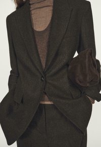 Kobieta ubrana w ciemnobrązową, dopasowaną marynarkę i spodnie, pod które ma na sobie brązowy sweter z golfem, trzyma ciemnobrązową torebkę z zamszu.