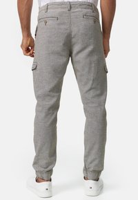 Man in grijze, omgeslagen cargobroek en witte sneakers, staand met ontspannen handen langs de zijkanten, gezien van achteren.