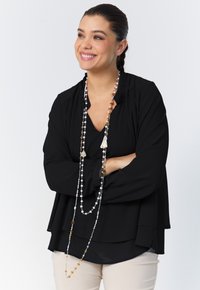 Black blouse with layered design, pleated neckline, and long beaded necklace featuring various colors and shapes. Beige pants below.