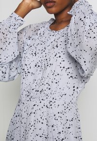 Hellgraues Kleid mit schwarzem Spritzmuster. Verfügt über Puffärmel, einen Knopfverschluss und eine geraffte Taille für eine figurschmeichelnde Silhouette.