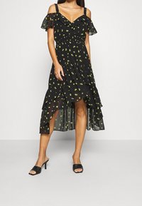 Frau trägt ein schwarzes Kleid mit kalten Schultern, gelbem Blumenmuster und asymmetrischem Volantsaum, kombiniert mit schwarzen High Heels.