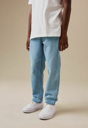 Kind trägt hellblaue Jeans, weiße Sneakers und ein weißes T-Shirt und steht vor einem einfarbigen beigen Hintergrund.