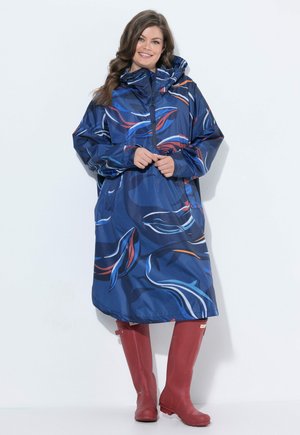 Mujer sonriente que lleva un impermeable azul con estampado y capucha y botas de goma rojas, posando frente a un fondo blanco liso.
