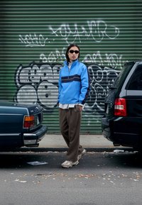 Μπλε αδιάβροχο με μαύρη ρίγα, φορεμένο πάνω από ένα ανοιχτόριγχο πουκάμισο και καφέ πλισέ παντελόνια. Γκρι αθλητικά παπούτσια, αστικό φόντο με γκράφιτι.