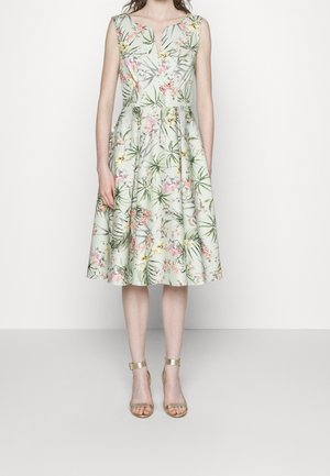 Frau trägt ein ärmelloses, knielanges hellgrünes Kleid mit Blumen- und Blattmustern, kombiniert mit goldfarbenen offenen Sandalen mit Knöchelriemen.