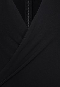 Černý textilní oděv s designem do zavinování. Vyznačuje se hladkou strukturou a viditelným zipem na zádech, čímž vytváří minimalistický vzhled.