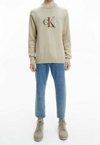 Pull beige en maille avec un col rond et un logo brodé marron, associé à un jean droit bleu clair et des chaussures à lacets grises.