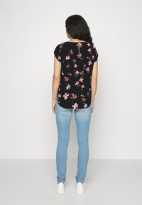 Blusa nera a fiori con maniche corte, caratterizzata da motivi floreali rosa e un dettaglio a chiave sulla schiena, abbinata a jeans azzurri chiari e sneakers bianche.