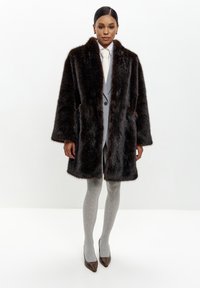 Brun faux fur frakke med bred krave, lange ærmer, lomme-lapper og en afslappet pasform, parret med grå ribbede strømpebukser og brune hæle.