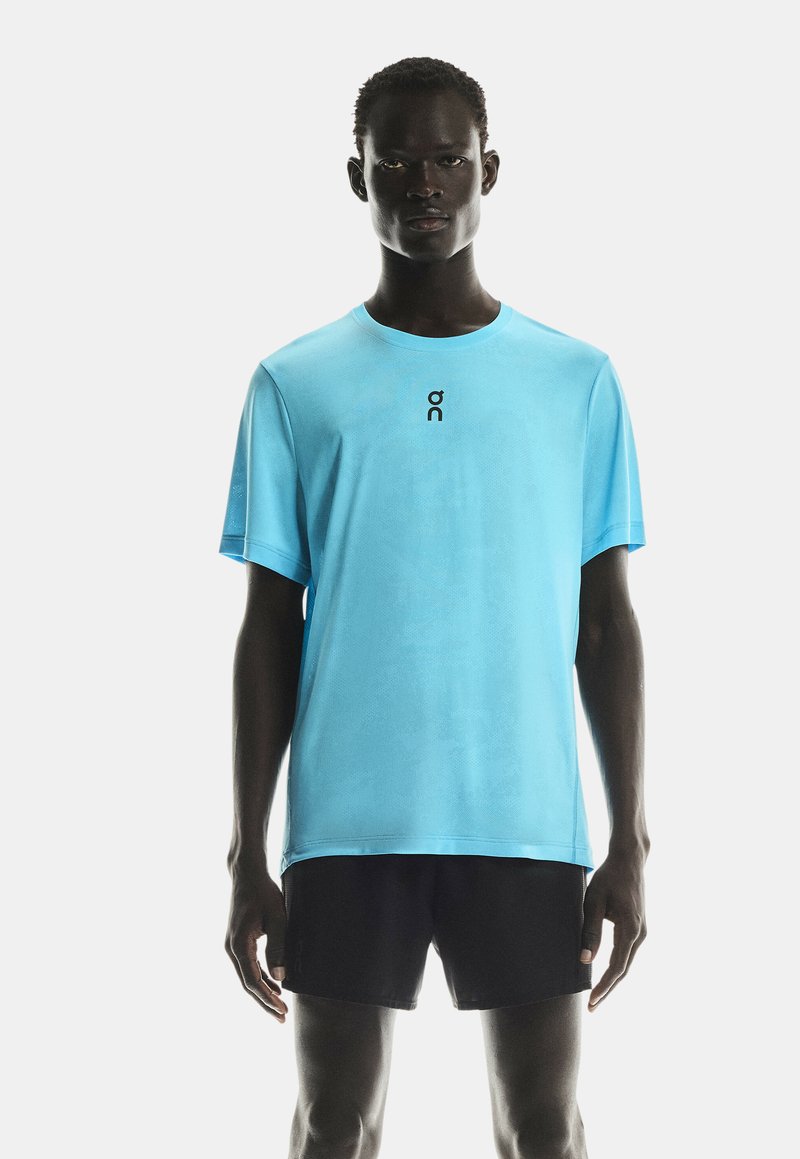 Model masculin îmbrăcat cu un tricou scurt din bumbac de culoare albastră deschis, cu un mic logo negru pe piept și pantaloni scurți sport negri, stând în fața unui fundal alb.