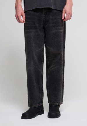 Persona che indossa jeans neri larghi e lavati e stivaletti neri, in piedi davanti a uno sfondo neutro.