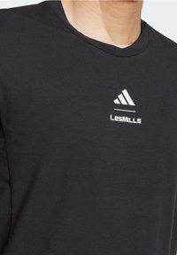 Sort T-shirt med hvid Adidas-logo og "LesMILLS" tekst på venstre bryst, båret af en person vist fra halsen og ned.