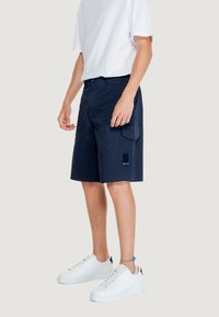 Pantaloni cargo blu con tasca e dettaglio del logo, realizzati in tessuto leggero. Abbinati a sneakers bianche e una maglietta bianca semplice.