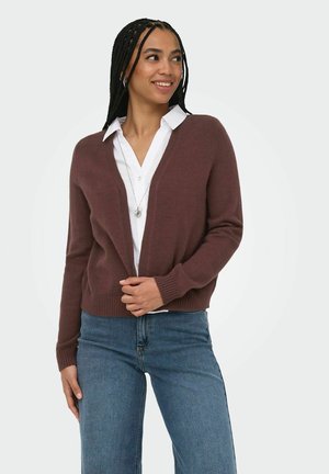 Donna che indossa un cardigan marrone sopra una camicia bianca e jeans blu, sorridente e guardando di lato, in piedi davanti a uno sfondo uniforme.