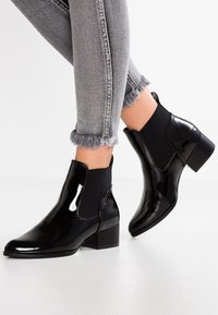 Bottines noires en cuir verni avec panneaux latéraux élastiques et talon carré court, associées à un jean gris déchiré avec des revers effilochés.
