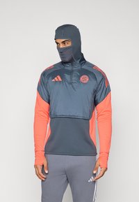 Grå og koralfarvet atletisk trøje med hætte, der har en halvdels lynlås-krave, tekstureret stof og Bayern München-logo på brystet. Grå bukser nedenunder.