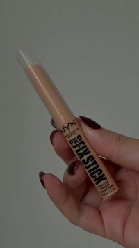 Mano con unghie di colore rosso scuro che tiene un correttore NYX Professional Makeup Pro Fix Stick con tappo trasparente su sfondo grigio.