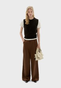 Zwarte gebreide mouwloze top over een wit shirt, gecombineerd met brede bruine broeken. Houdt een kleine beige tas vast. Eenvoudig, strak ontwerp.