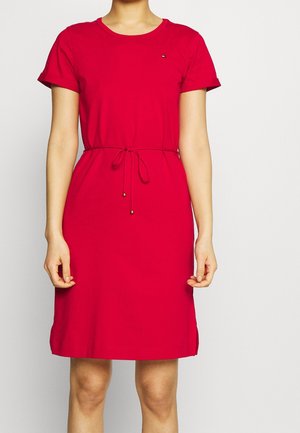 Kvinde iført en rød kjole til knæet med korte ærmer og bælte i taljen, stående mod en ensfarvet lys baggrund.